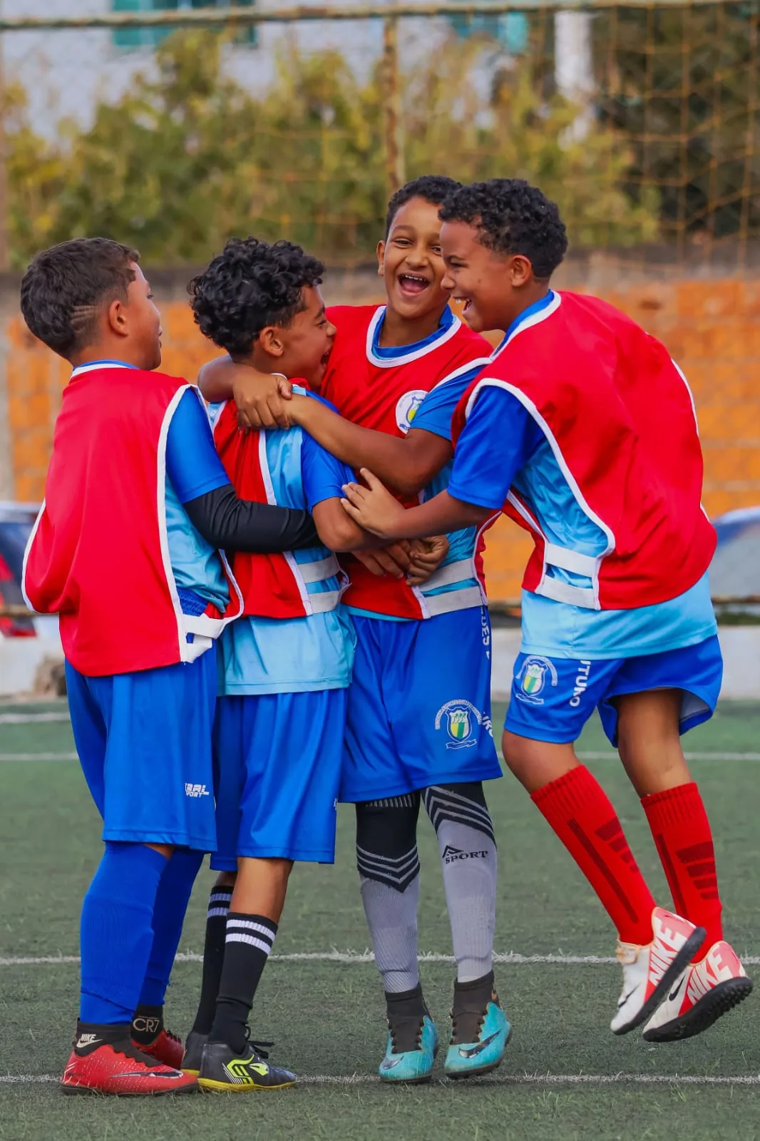 Quatro alunos de colete vermelho e azul se abraçando para comemorar um gol